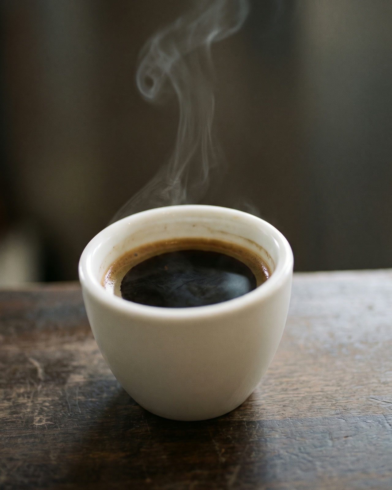 A small white cup of black coffee on a dark counter at night with a thin wisp of steam